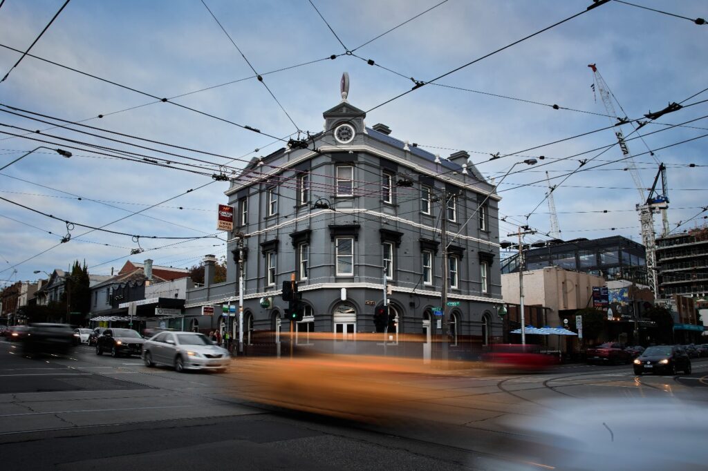 The Swan Hotel exterior at dusk, a heritage corner pub in Melbourne with tram wires overhead and light trails from passing traffic.